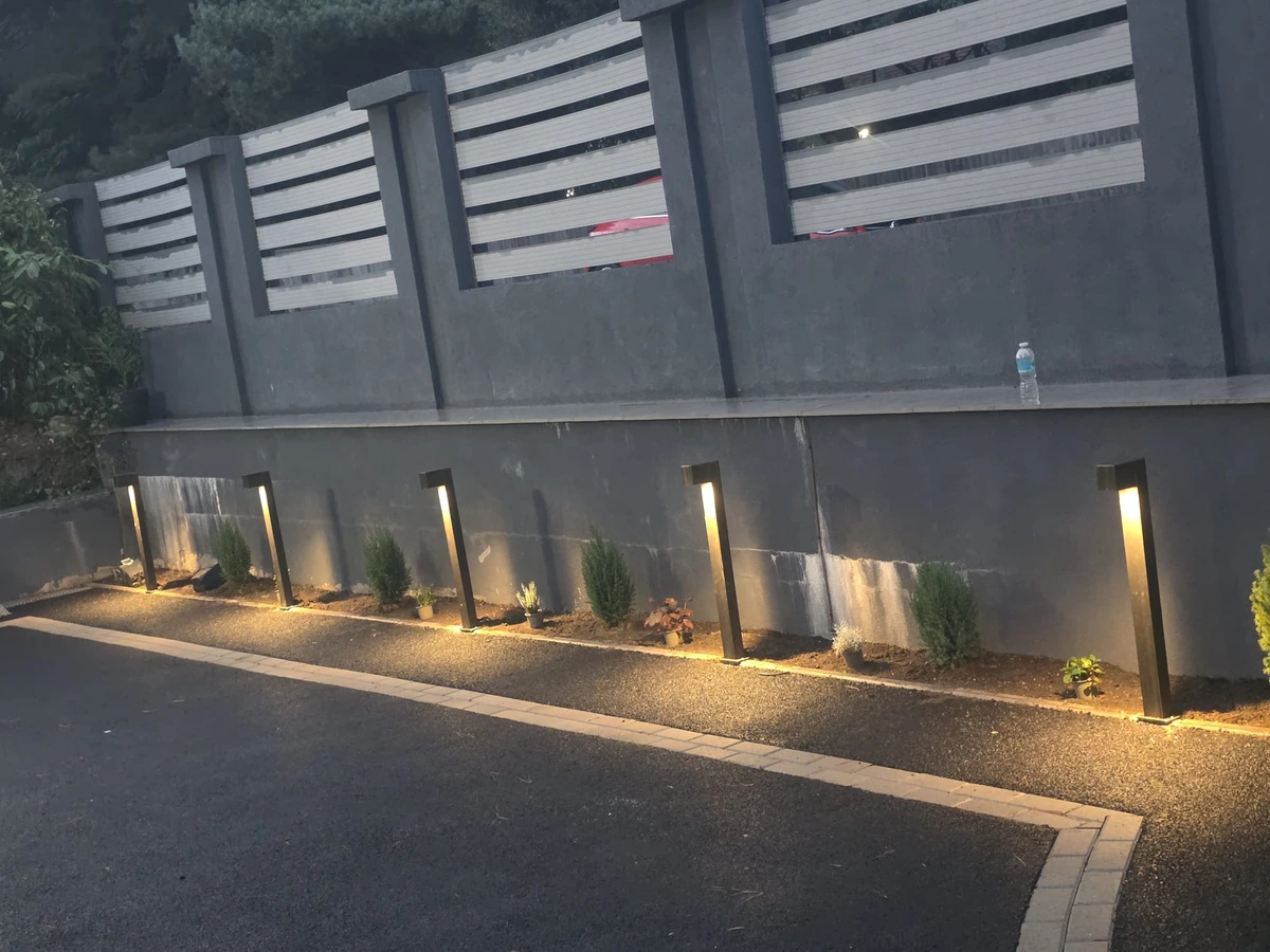 Row of outdoor bollard lights illuminating a driveway beside a slatted boundary fence